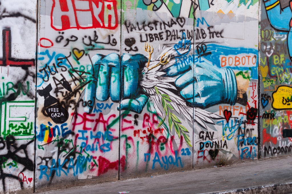 Un tratto del muro di separazione a Betlemme. Un graffito rappresenta due mani vestite con due guanti azzurri. In una delle due mani una colomba viene stretta e schiacciata in un pugno, mentre un dito dell'altra mano la spinge all'interno del pugno. Diverse altre scritte circondano il graffito.