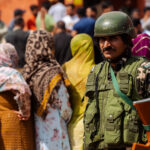 Un soldato in Kashmir presidia i seggi. Sullo sfondo una fila di donne in attesa di votare.