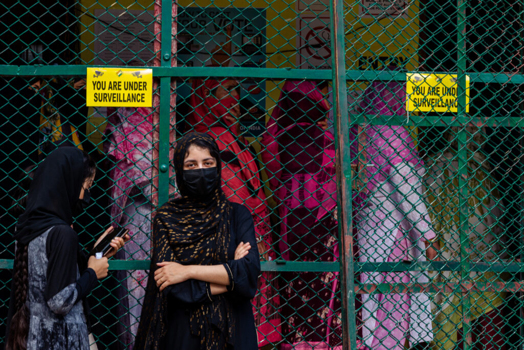 Donne in fila ad un seggio elettorale nel distretto di Baramulla in Kashmir. Una donna guarda in camera, mentre altre donne sono in fila dietro una grata di ferro. Un cartello recita "siete sotto sorveglianza".