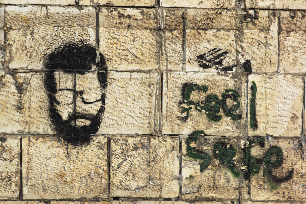 A wall in the city of Ramallah with a stencil depicting the stylized face of a Palestinian martyr. His face has a beard, mustache and glasses. A writing on the wall, not easily readable, seems to say "feel free".