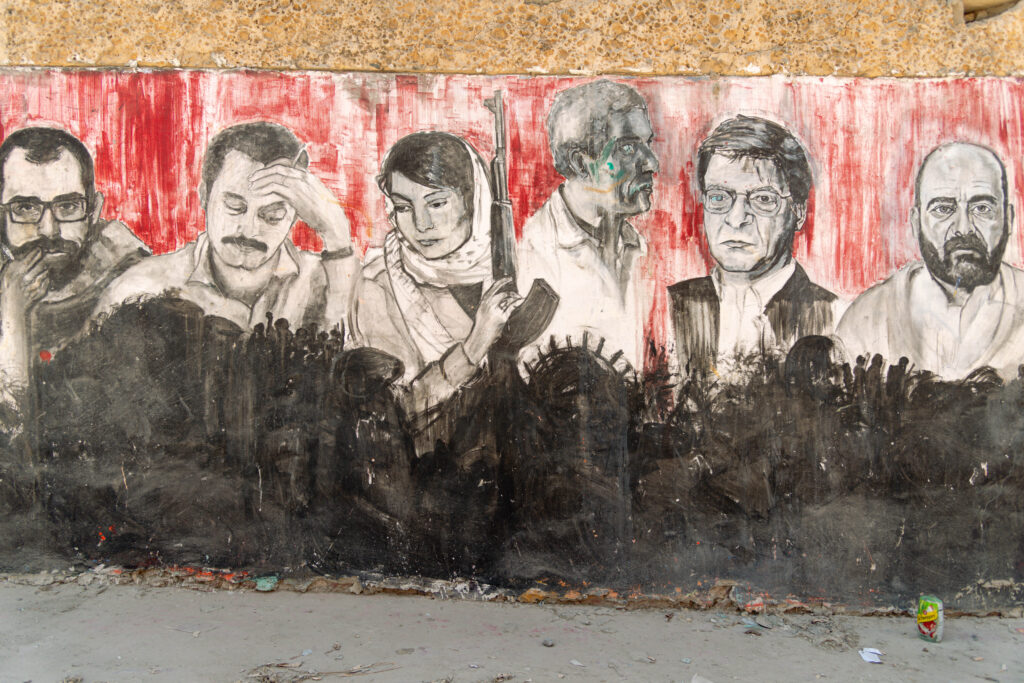 A mural in the Jenin refugee camp, outside the Freedom Theatre, one of the most interesting cultural and resistance realities of Palestinian society. The mural represents some of the most iconic personalities of Palestinian history, including the writer Ghassam Kanafani and the freedom fighter and activist Leila Khaled. The drawings are in black and white, while the background is red. At the base of the mural a black silhouette recalls crowds of Palestinians and the head of Handala.
