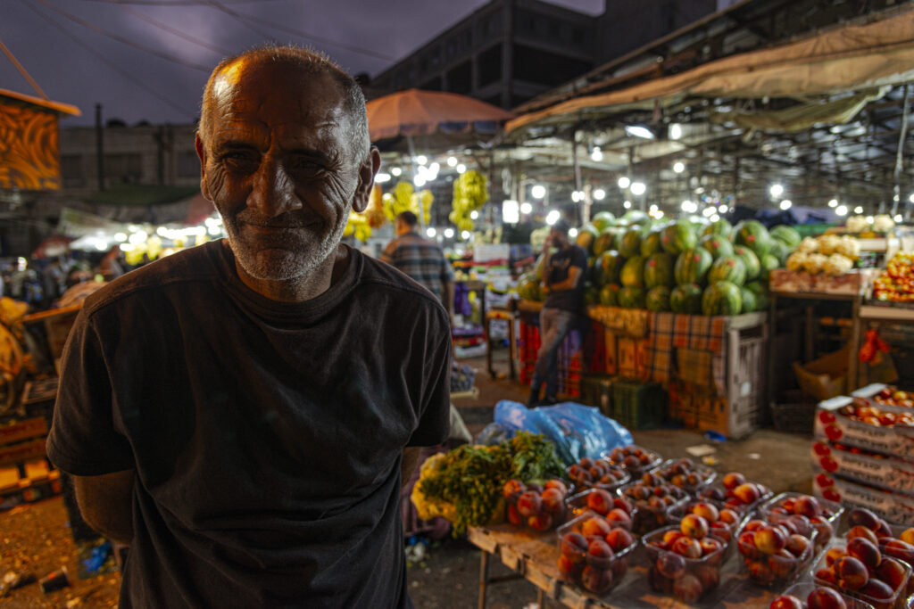 Ramallah, Palestine - An elderly fruit and vegetable vendor poses for a nighttime photo. Behind him the colorful stalls of the farmers' market.