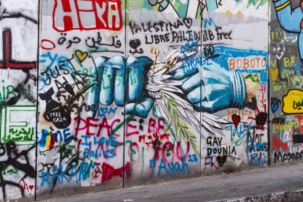 A section of the separation wall in Bethlehem. A graffiti represents two hands dressed in two blue gloves. In one of the hands a dove is held and crushed in a fist, while a finger of the other hand pushes it into the fist. Several other writings surround the graffito.