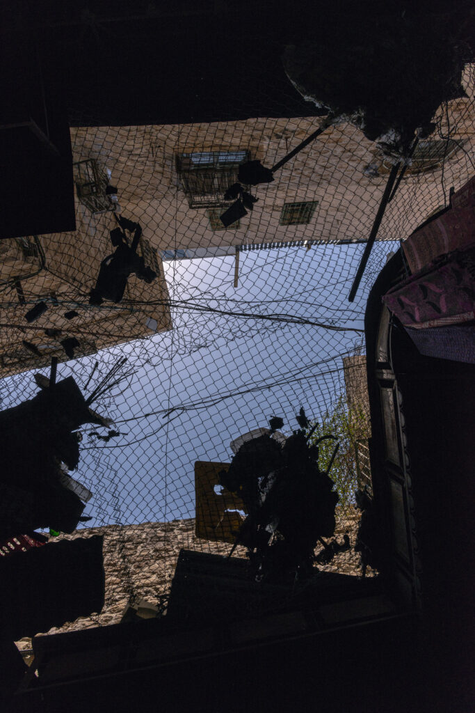 Una foto dal basso in una delle vie del mercato di Hebron mostra una rete che separa il piano strada dai piani superiori, dove le case sono state occupate dai coloni israeliani. I coloni israeliani lanciano rifiuti sulla strada e sugli abitanti palestinesi di Hebron. La rete è stata montata per riparare gli abitanti dalla pioggia di rifiuti. Spesso i rifiuti rimangono lì per anni. La vista del cielo è interrotta da cumuli di rifiuti bloccati dalla rete