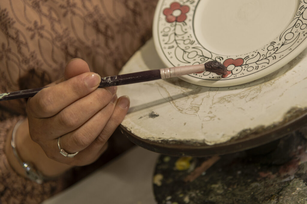 The hand of one of the craftswomen holds a paintbrush as she decorates one of the plates with a floral pattern.
