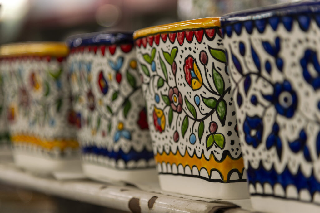 Some pottery is ready for sale on a shelf in the store adjacent to the workshop. The ceramics have floral patterns. In the foreground is a blue-decorated pottery; the second, in focus, is decorated with yellow and red flowers.