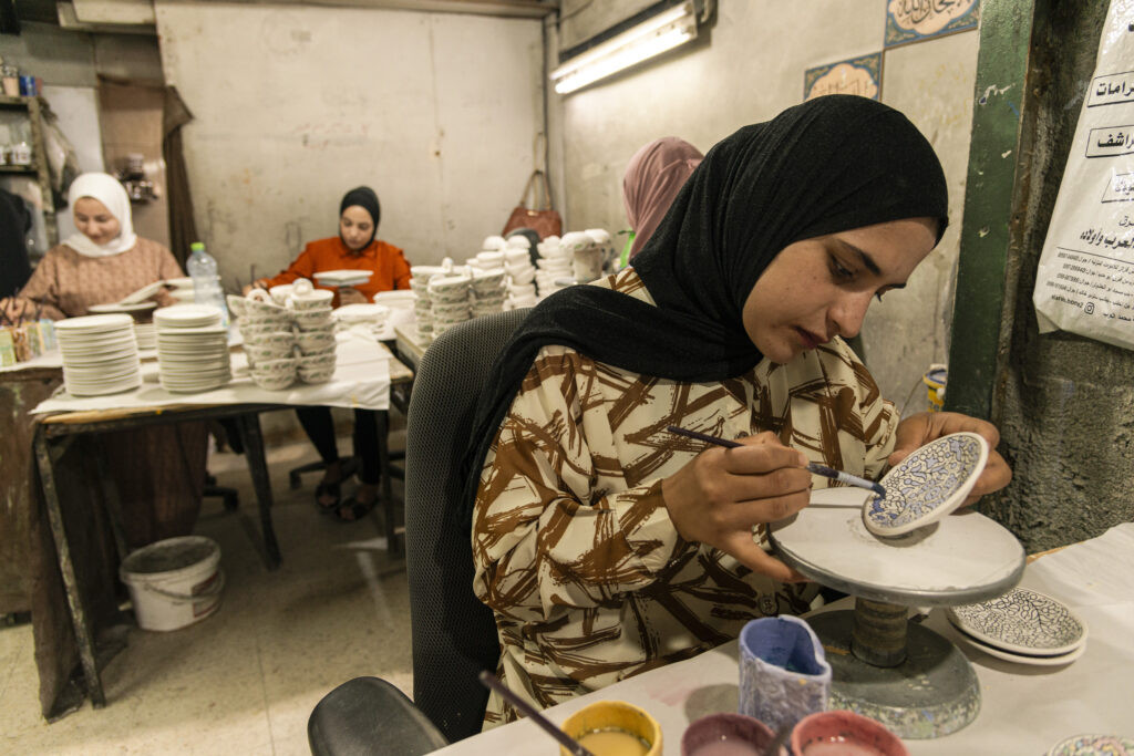 A girl decorates a ceramic plate. Behind her, in the workshop, other girls work behind a stack of ceramic plates.