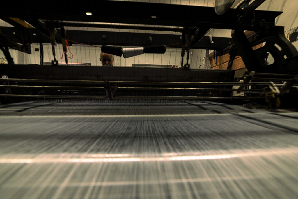Palestine's last Kufiyah factory in Hebron. An elderly craftsman is photographed from behind the loom as he works. In the foreground are the white threads of the loom, still not woven into the typical decorations that characterize Kufiyah.