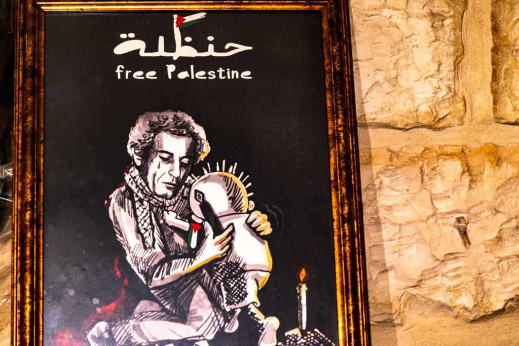 Inside the Handala Café in Ramallah, a poster inside a frame depicts Naji al-Ali, the author of Handala, who embraces his character, as if embracing all Palestinian children. A tear runs down the face of Naji al-Ali, who in one hand holds a pen to draw. Above them a sign reads "free palestine". On Handala's shoulder hangs a Palestinian flag. A candle illuminates two characters.