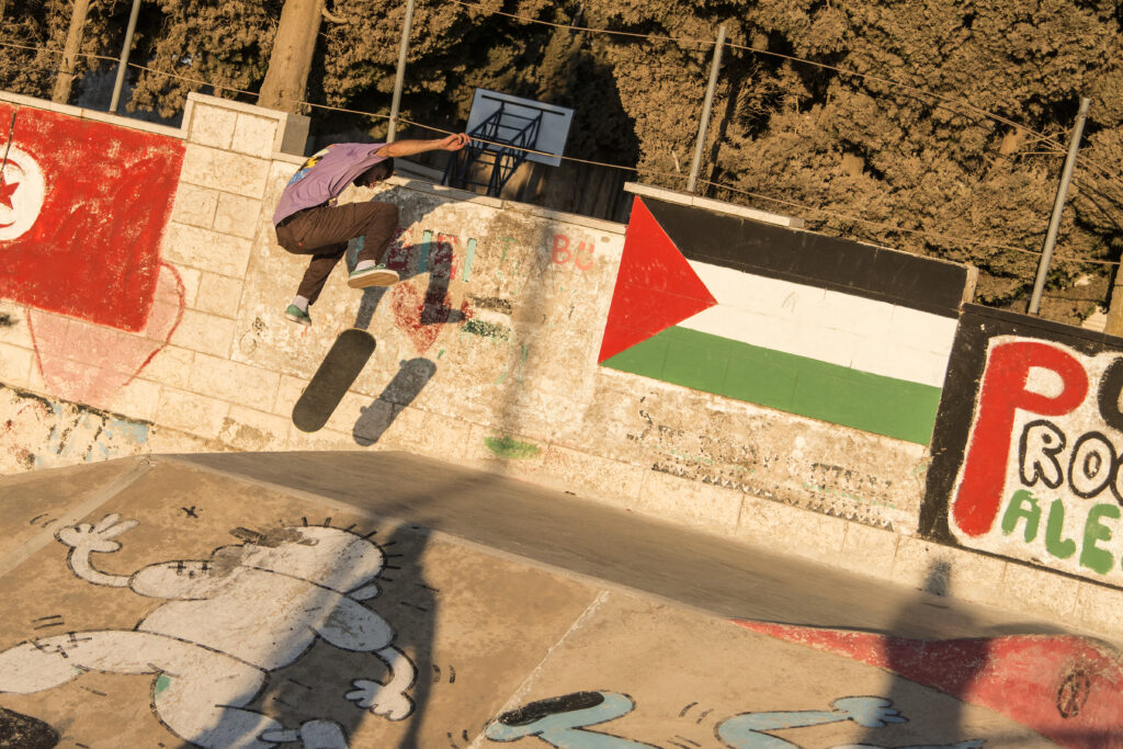 A view of the skatepark near Nablus. A boy jumps over a ramp on his skateboard. The ramp is painted with a Palestinian flag and graffiti by Handala.