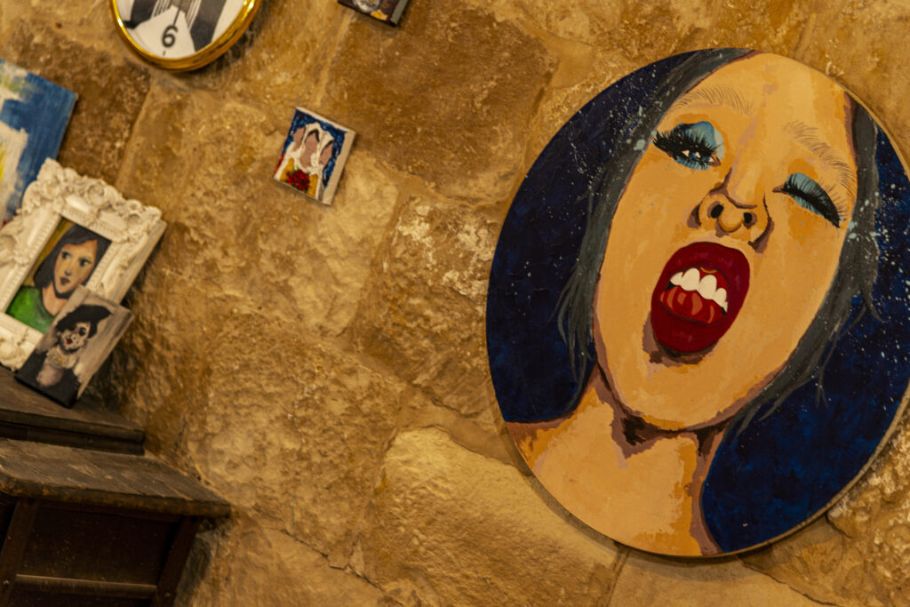 A wall with several paintings in the coffee lab restored as part of The Yalla Project. There are several paintings on the stone wall. One in particular takes up a large part of the photo, with a woman's face depicted on an oval canvas.