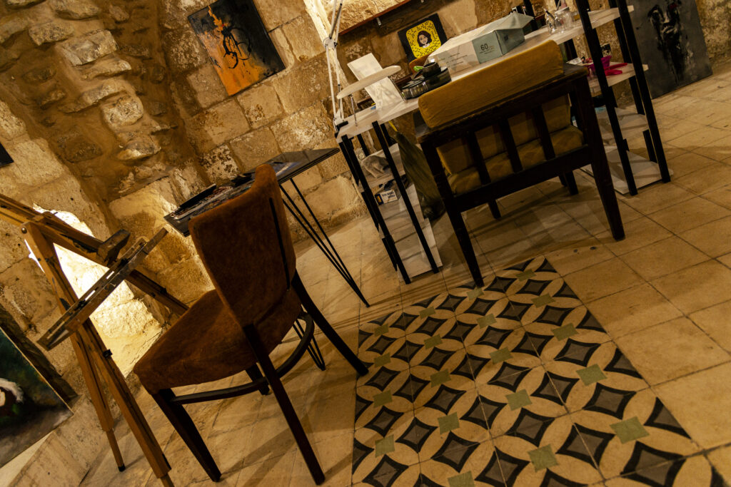 A glimpse of the workshop of one of the buildings restored as part of The Yalla Project. Inside, dozens of paintings. In the photo, you can see a chair in front of an easel and some paintings on the rock walls. The floor is tiled with a typical Palestinian architectural motif.