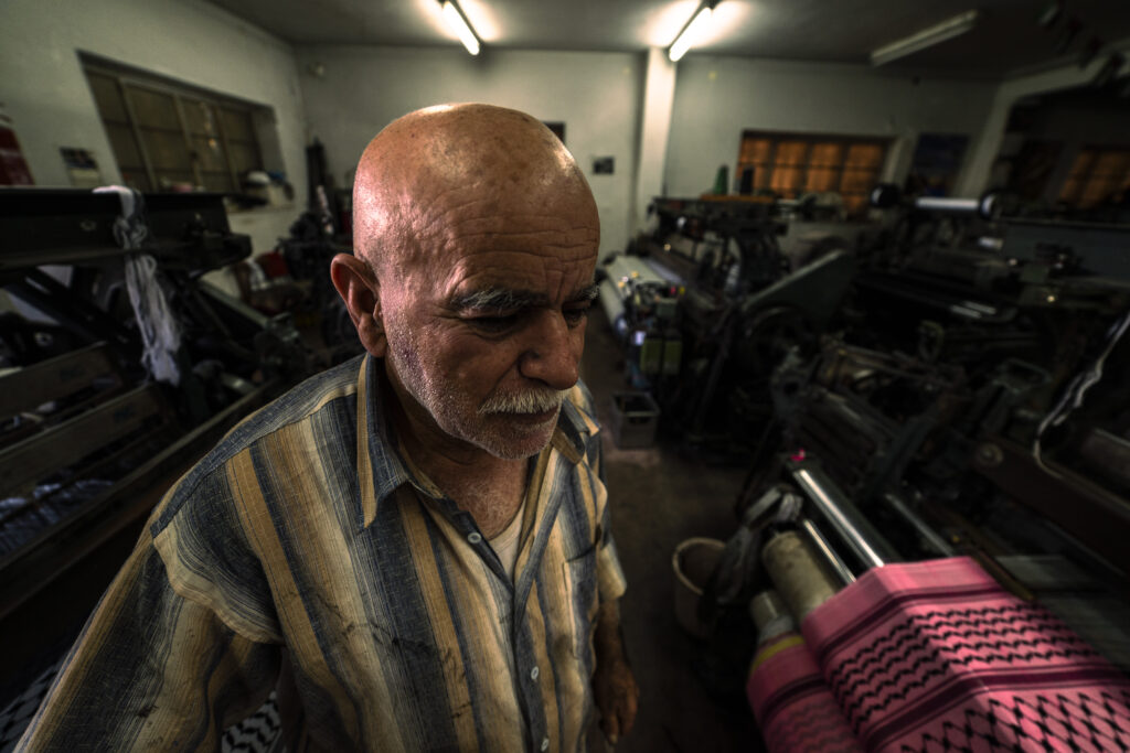 The last Kufiyah factory in Hebron, Palestine. An elderly craftsman stands in the foreground, looking down with a look that seems pensive. Behind him a few looms, inside a shed lit by halogen lamps.