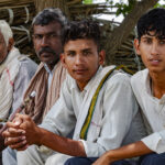 Quattro braccianti, appartenenti alla stessa famiglia posano guardando in camera. Sono seduti. e ripresi di taglio I due fratelli più giovani sono in primo piano e dietro di loro i loro parenti più anziani. Sono in pausa dal lavoro e si trovano al riparo dal sole, all'ombra di un albero.