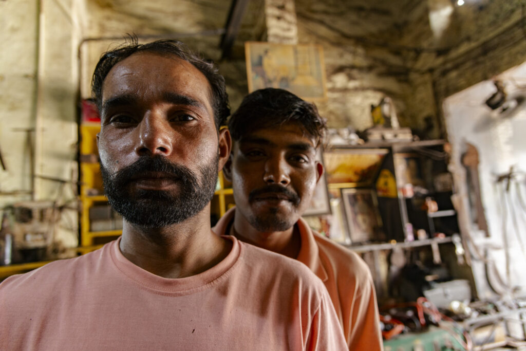 Two young carpenters pose inside the workshop where they work. The two young men are in the foreground while behind them on the wall hang various tools and work instruments.