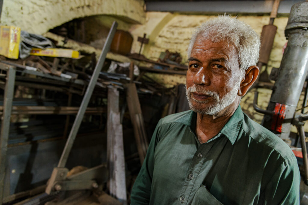An elderly carpenter is pictured inside his workshop, where he works with several other people.