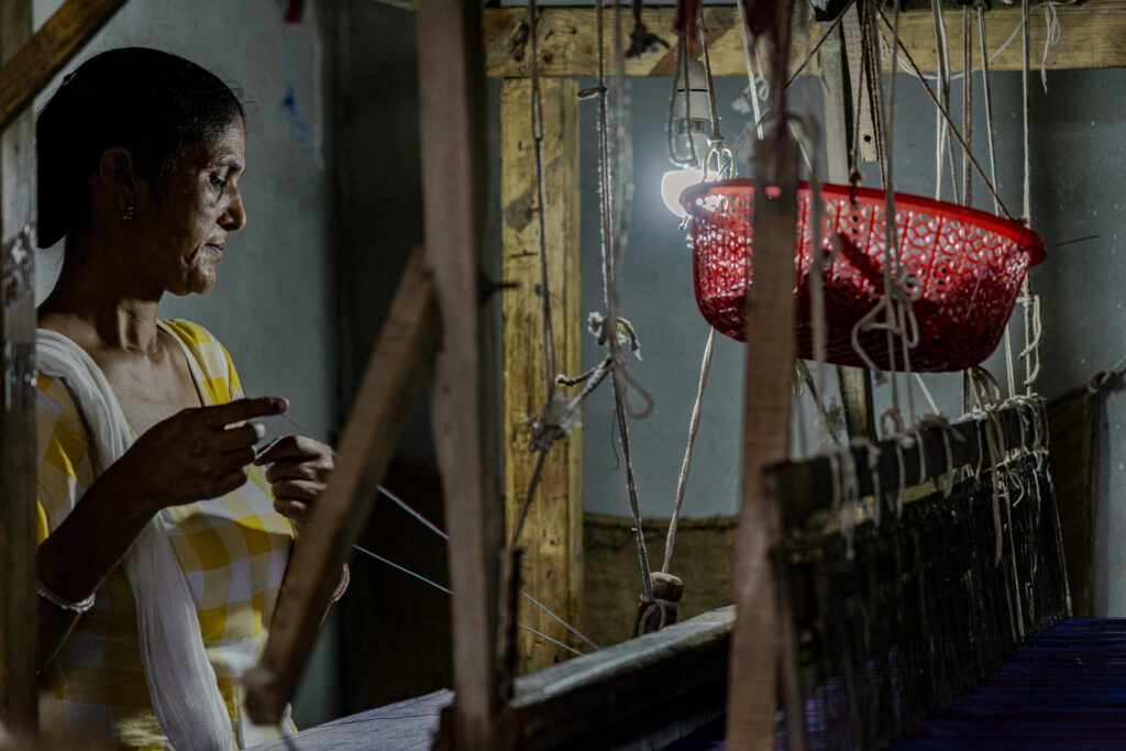 A woman works at a loom. In the picture, the woman is shot from the side. A light illuminates her face and loom, while the rest of the image is in half-light. The woman holds a thread in her hands as she prepares a decoration for the fabric she is making.