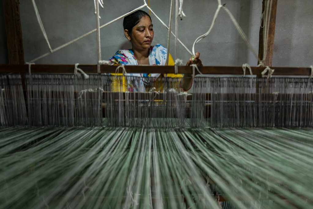 A woman works at a loom. In the photo, the woman is shot from the front. The threads of the loom can be seen in the foreground while she is working in the background.