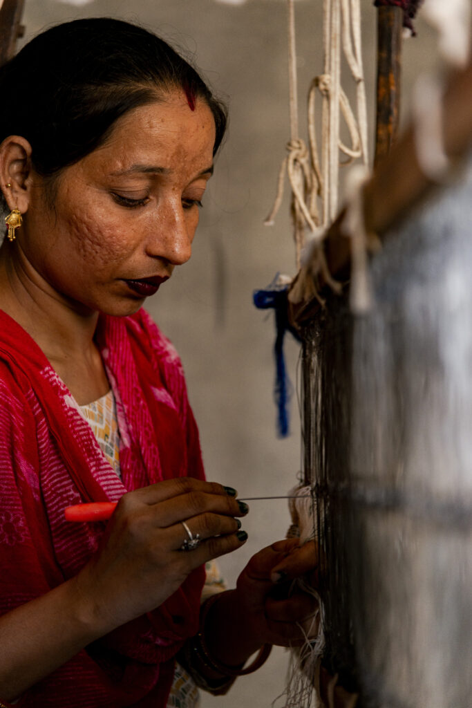 A school student prepares the loom for weaving. She is shot cutting. Her gaze is downward as she is focused on threading one of the hundreds of threads inside one of the hooks that will be used to alternately move the threads up and down to create the fabric