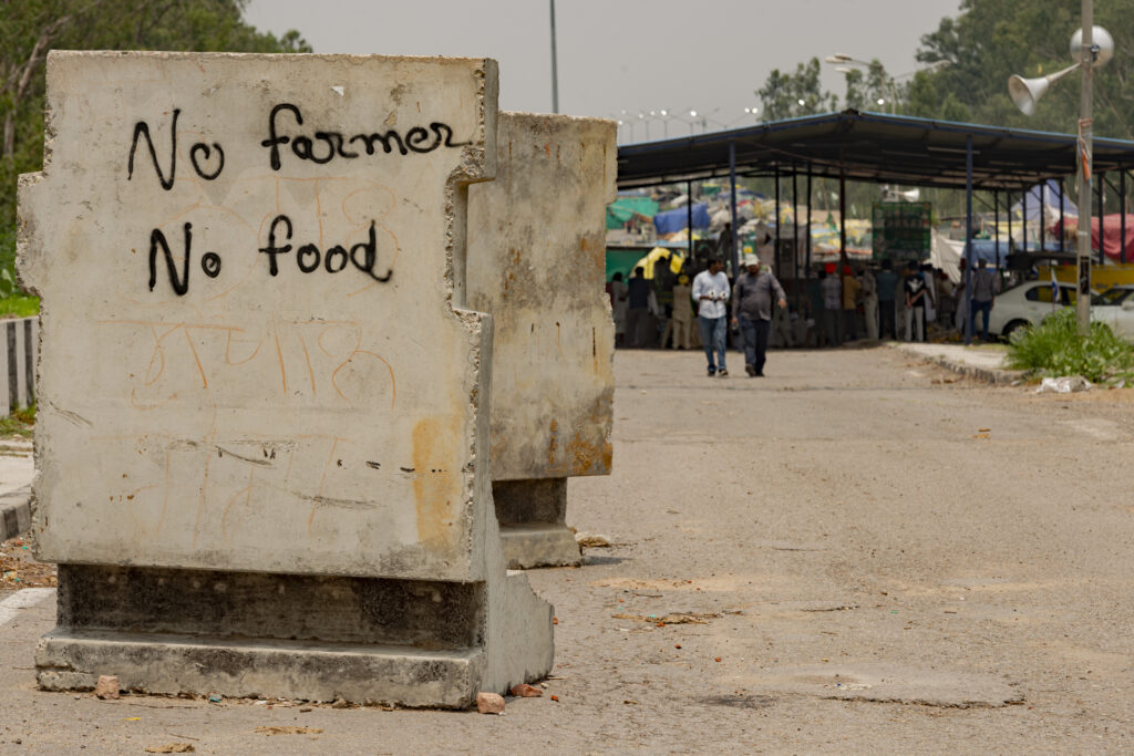 Una scritta su un jersey in cemento recita "no farmers no food" nel sito del sit-in permanente presso lo Shambhu Border, lungo l'autostrada che porta verso Delhi dal Punjab. Sullo sfondo agricoltori durante un comizio.